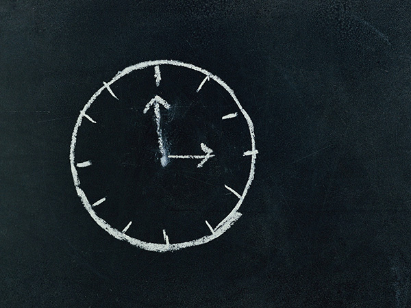 A simple drawing of an analog clock is shown, sketched with white chalk on a blackboard in a Catholic school classroom. The clock's hands are pointing at approximately 10:10.