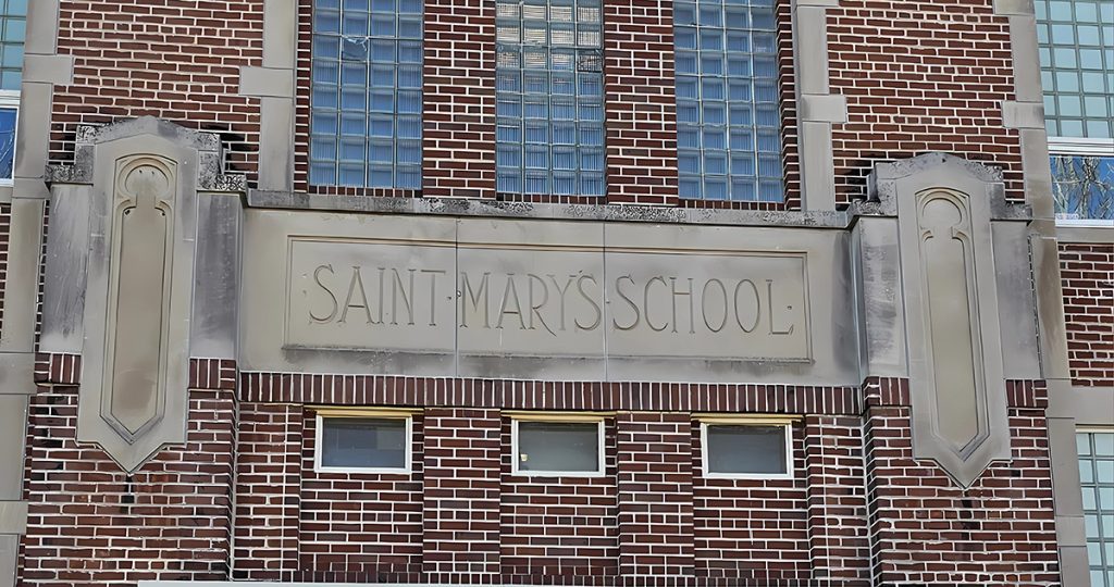 A brick building facade with large glass block windows and a stone sign reading "Saint Mary's Catholic School." The sign is flanked by decorative stone panels. Three small windows are below the sign, completing the building's traditional architectural style.