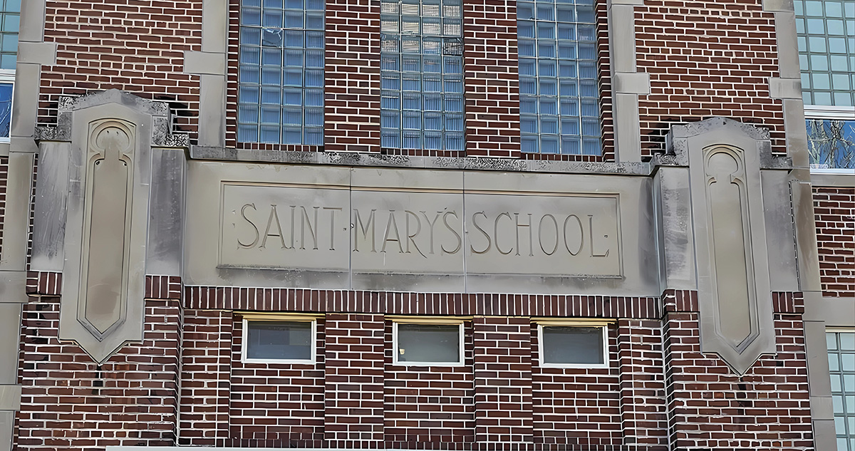 A brick building facade with large glass block windows and a stone sign reading "Saint Mary's Catholic School." The sign is flanked by decorative stone panels. Three small windows are below the sign, completing the building's traditional architectural style.