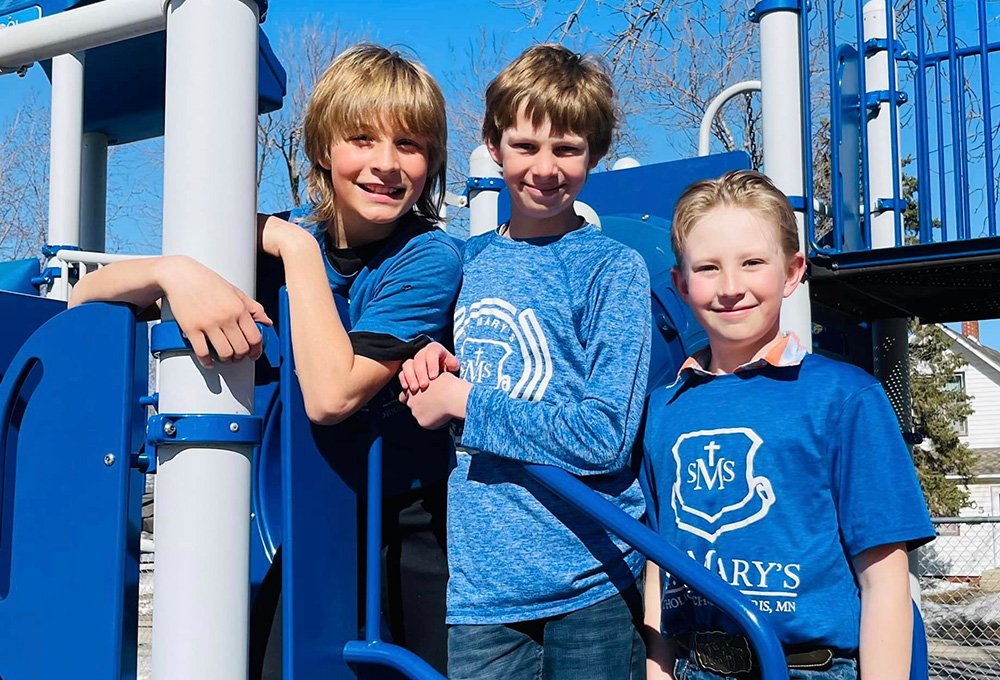 Three boys pose together, smiling at an outdoor playground. They are wearing blue shirts with "St. Mary's Catholic School" printed on them. The playground equipment and a clear blue sky are visible in the background.