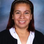 Yadira Lara Guerrero, a woman with shoulder-length auburn hair and a black headband, smiles at the camera. She wears a white shirt with vertical stripes and a black cardigan. The background is a gradient of blue and gray tones.