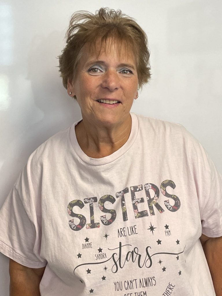 A woman with short, light brown hair is smiling at the camera. She is wearing a light pink T-shirt with the word "SISTERS" on it, decorated with floral patterns. The shirt also has a saying about sisters beneath the word. The background is plain white. This feels like a perfect tribute to Pam Gustafson's enduring message about sisterhood.