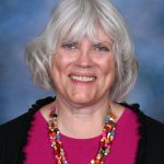 A smiling woman with shoulder-length gray hair and bangs, Mary Henrikson is wearing a colorful bead necklace and a pink top. She has a black cardigan over her top. The background is a gradient of blue shades, creating a soft and pleasant backdrop for the portrait.