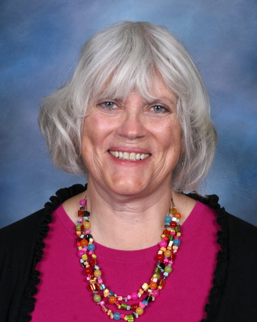 A smiling woman with shoulder-length gray hair and bangs, Mary Henrikson is wearing a colorful bead necklace and a pink top. She has a black cardigan over her top. The background is a gradient of blue shades, creating a soft and pleasant backdrop for the portrait.