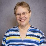 Linda Hodgson, a woman with short, grayish hair, is smiling at the camera. She is wearing glasses and a striped shirt with shades of blue, white, and yellow against a plain gray backdrop.