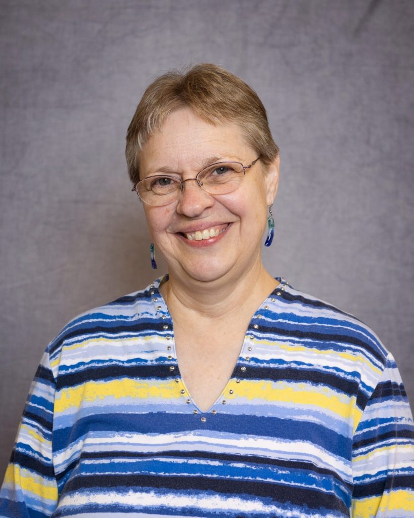 Linda Hodgson, a woman with short, grayish hair, is smiling at the camera. She is wearing glasses and a striped shirt with shades of blue, white, and yellow against a plain gray backdrop.