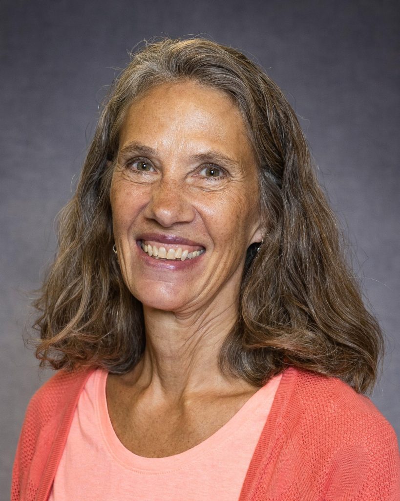 ViAnn Larson, a woman with wavy, shoulder-length gray hair, is smiling at the camera. She is wearing a coral pink top and a matching cardigan. The background is a plain gray color.
