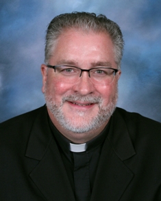 Fr. Todd Schneider, a middle-aged man with gray hair and a beard, wearing glasses and a black clerical shirt with a white collar, smiles at the camera. The background is a gradient of blue and purple tones.