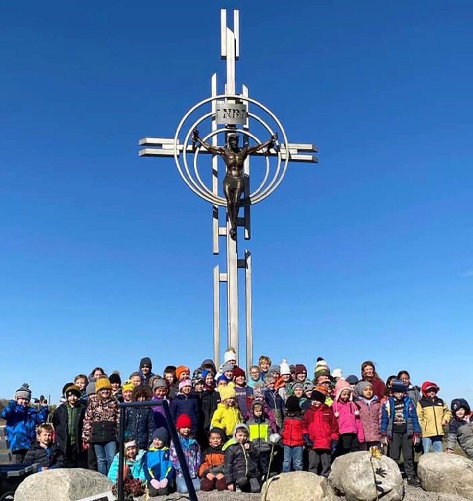 A large group of children in colorful winter clothing stands in front of a tall cross with a statue of Jesus Christ, set against a clear blue sky. Some children are seated on large rocks, and they all appear to be posing for a group photo.