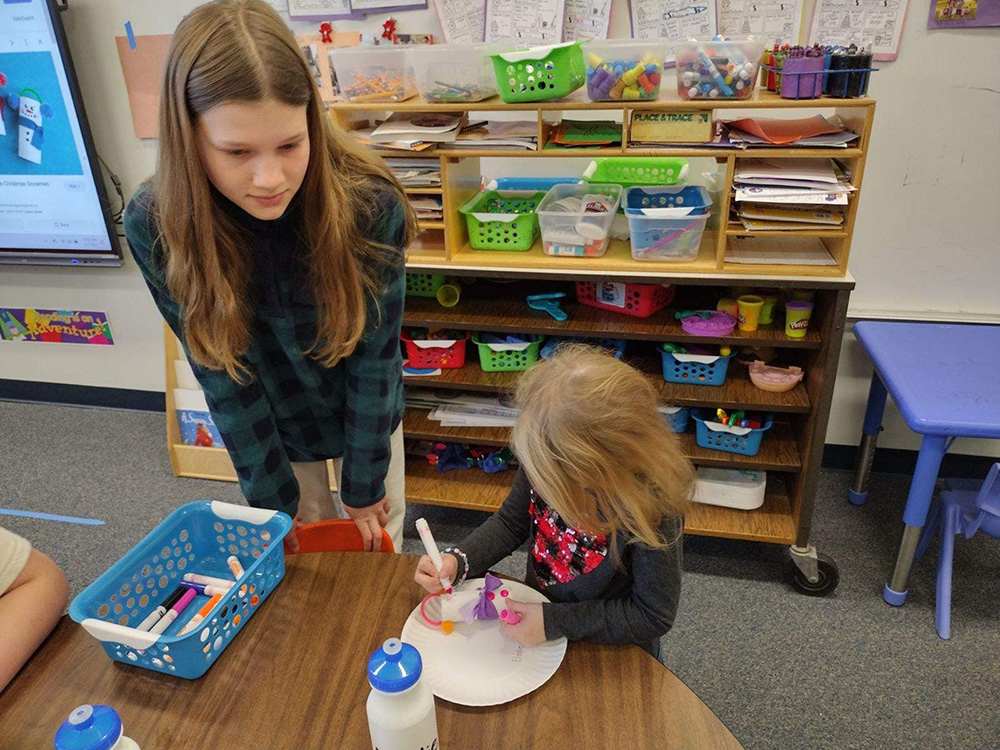 A young girl stands and observes while a younger child colors on a paper plate with markers in a classroom. The table is scattered with other markers and a water bottle. Shelves behind them contain various classroom supplies like bins, papers, and toys.