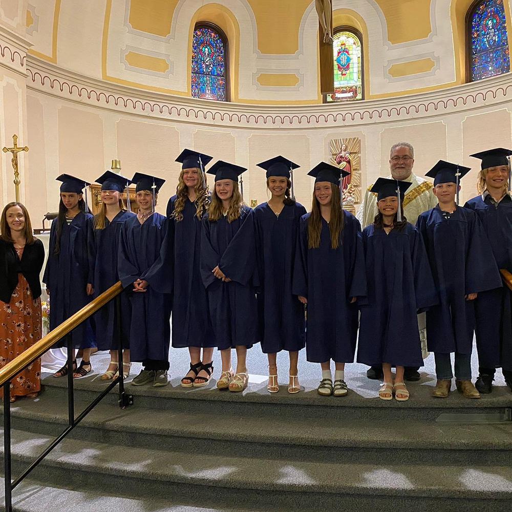A group of graduates wearing navy blue caps and gowns stand on steps inside a church, posed for a photo. There are two adults, one at each end of the group. Stained glass windows and religious symbols are visible in the background.
