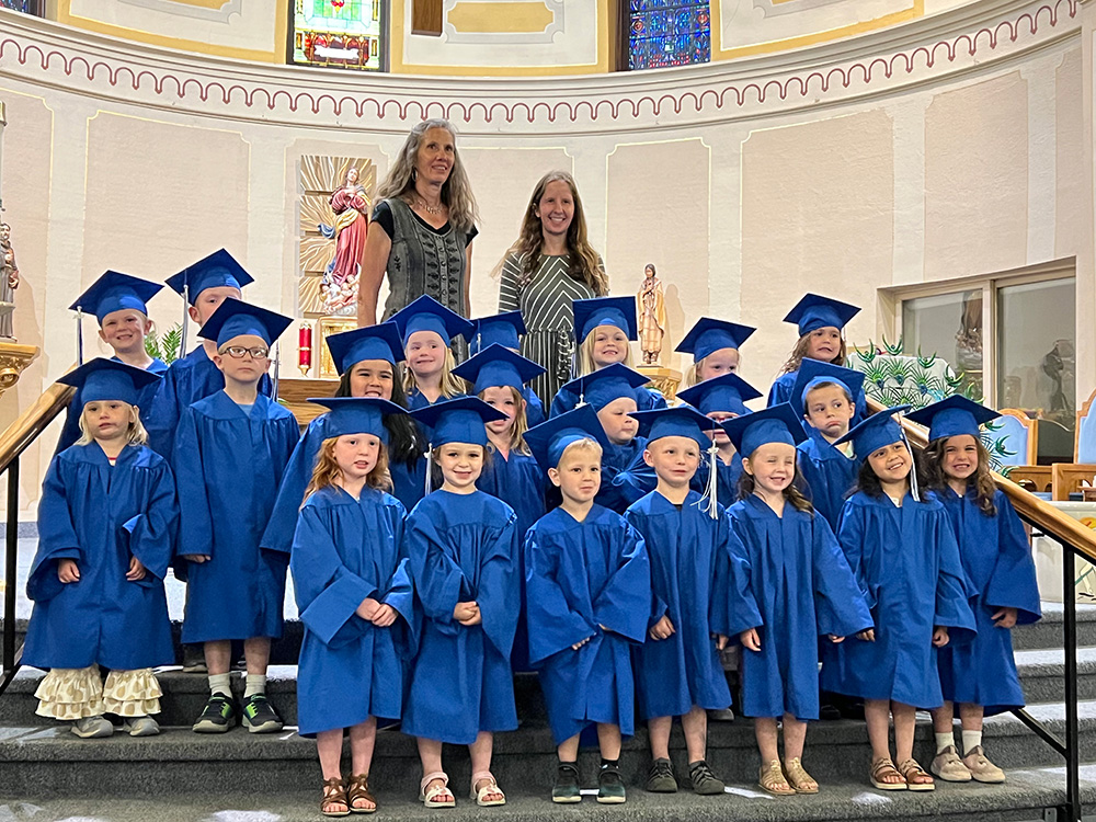 A group of young children, dressed in blue graduation gowns and caps, stands on the church steps, smiling. Two adults stand behind them, also smiling. The scene reflects a celebration of their early academic achievements against a backdrop of religious statues and stained glass windows.