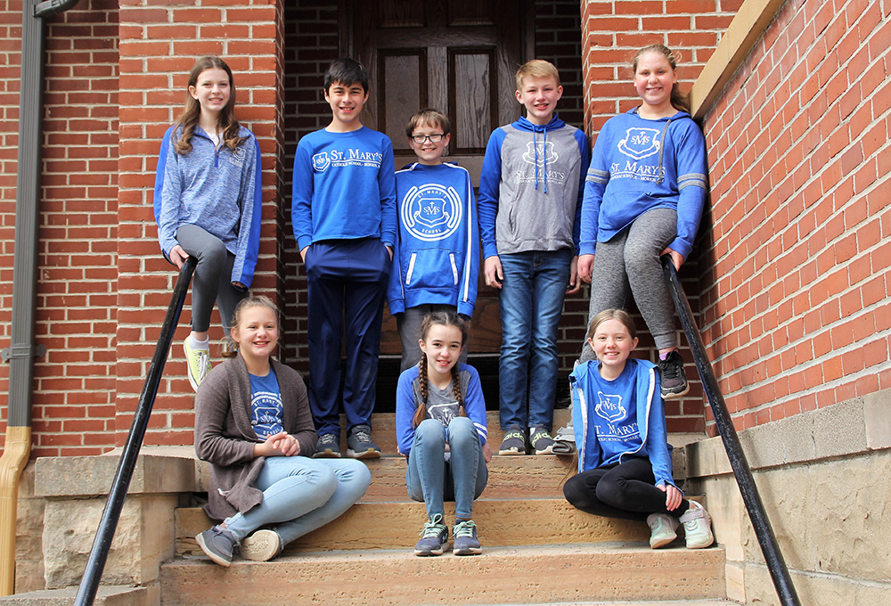 A group of eight children pose on the steps of a brick building. They are smiling and dressed casually, most wearing blue and gray clothes, with several in matching school-branded apparel. Four children are standing at the top, and four are sitting on the steps, but unfortunately, the provided description "About" is too vague to identify specific SEO keywords.