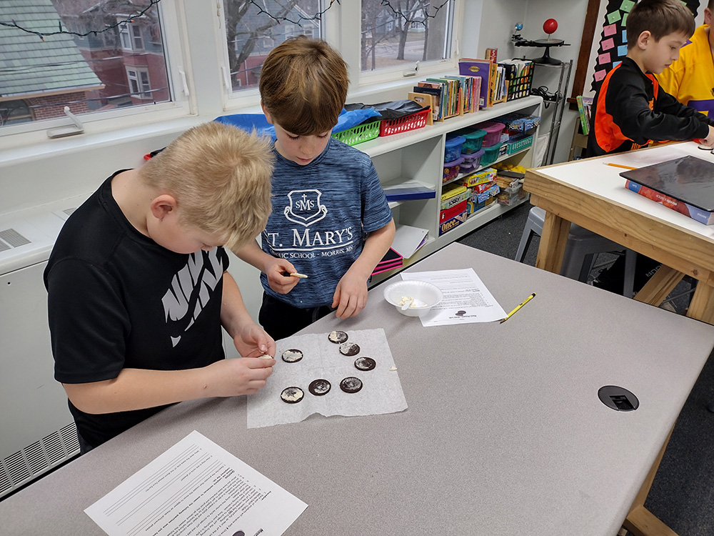 Two young boys are standing at a classroom table, examining Oreo cookies on a paper towel. One is peeling the cookies apart while the other looks on. There are papers and books on the table and shelves in the background. Another child is visible off to the side.
