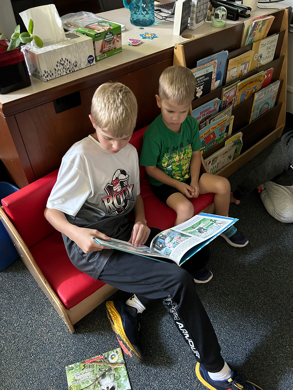 Two young children sit together on a small red couch. The older child, dressed in a white sports shirt and dark pants, reads a book to the younger child in a green shirt. Behind them is a bookshelf filled with a variety of colorful books.