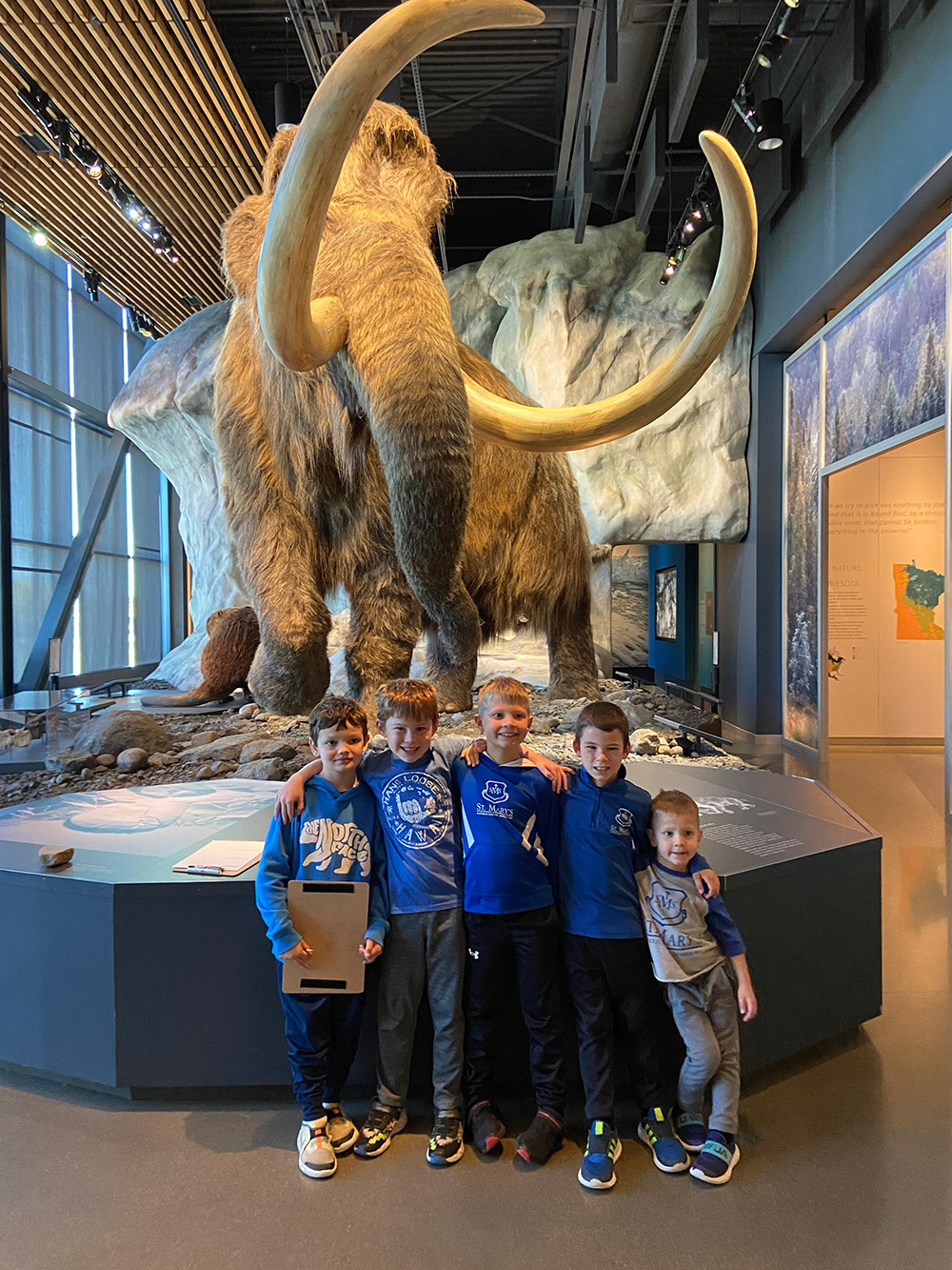 Five boys stand in front of a mammoth exhibit at a museum. They are smiling and posing for the photo, wearing casual clothes and shades of blue. The background features a large mammoth model and information displays with a rocky and snowy setting.