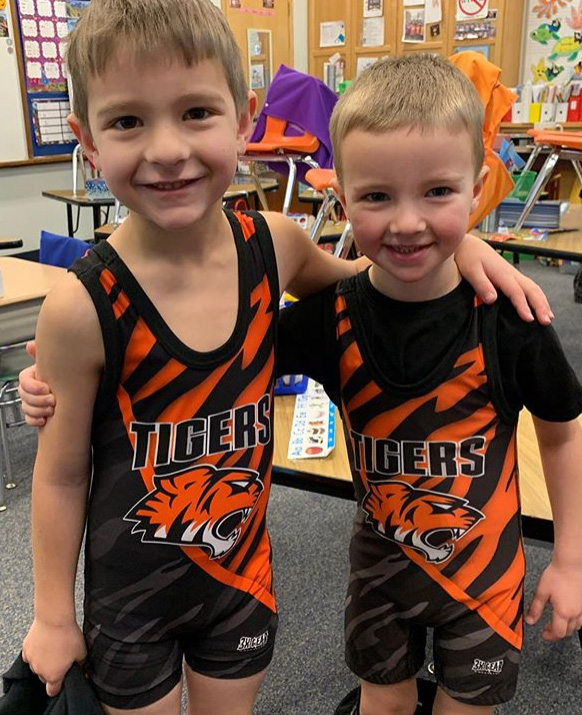 Two young boys smile while standing in a Pre-K classroom. They are wearing matching athletic uniforms with an orange and black tiger design and the word "TIGERS" written across the front. The classroom, part of a new educational program, has desks, chairs, and colorful decorations.