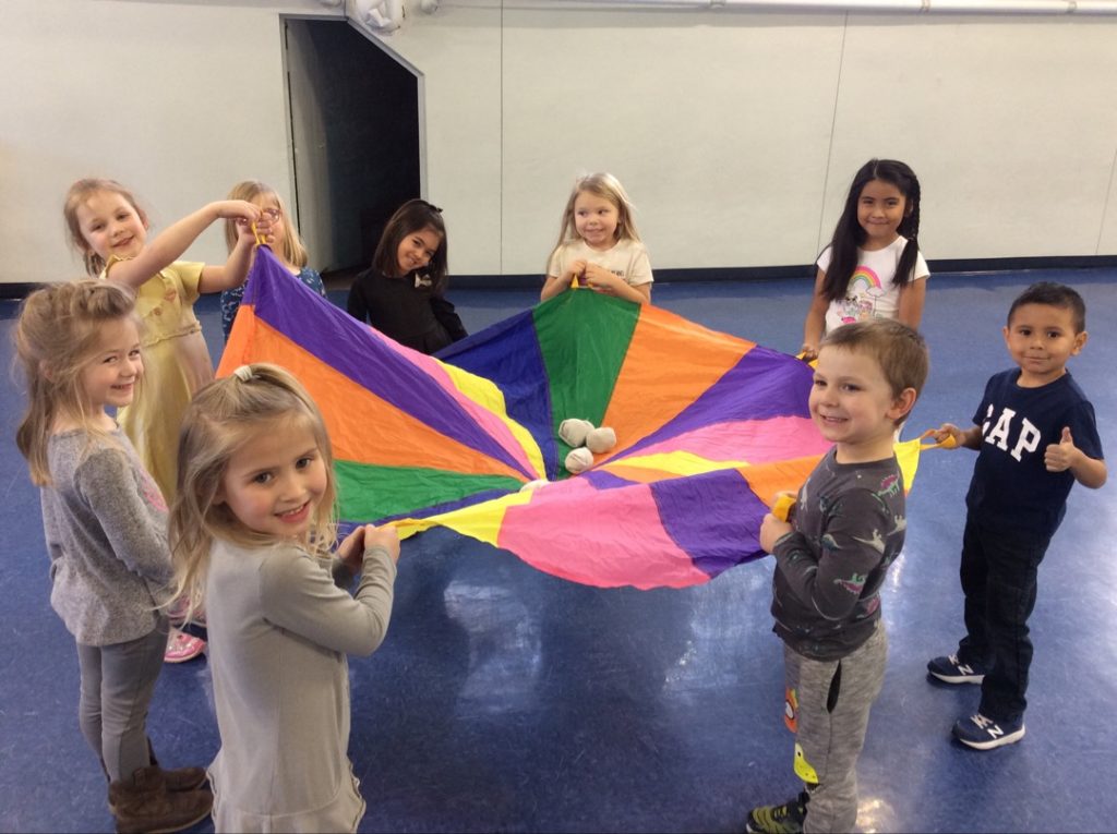 A group of young children stand in a circle holding a colorful parachute. They are smiling and appear to be enjoying an early education activity. Balls are placed on the parachute, fostering child development. The room has a plain wall and a blue floor.