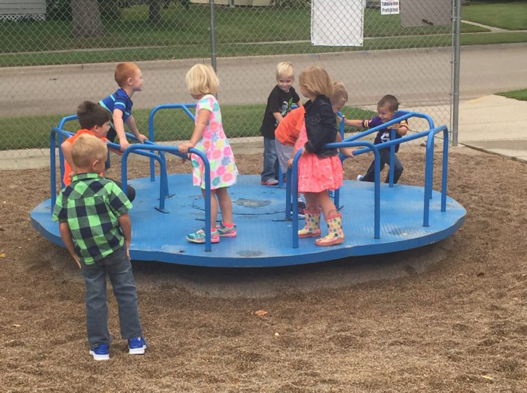Children in a Pre-K program play on a blue merry-go-round in a playground. Most of the kids are standing on the merry-go-round, while one child in a green plaid shirt stands nearby on the gravel surface. The playground is enclosed by a chain-link fence.