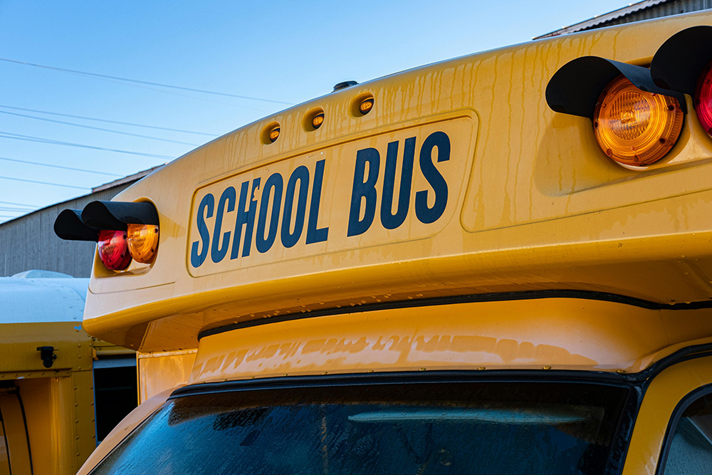A close-up view of the front of a yellow school bus with the words "SCHOOL BUS" prominently displayed above the windshield. The bus has red and amber lights on top, with condensation or frost visible due to cold weather. Route information can be critical during chilly mornings under a clear blue sky in the background.