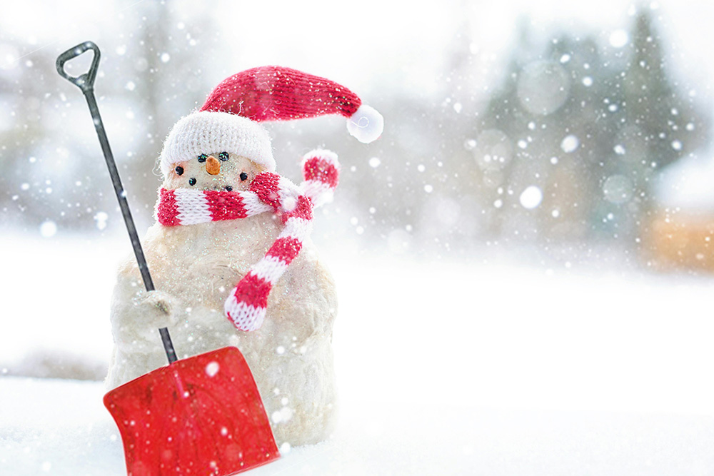 A snowman wearing a red and white striped scarf and a red Santa hat stands in a snowy landscape holding a red snow shovel. Snowflakes are falling around it, perfectly capturing the magic of a Snow Day. In the background, trees blend into the blurred snowy scene.