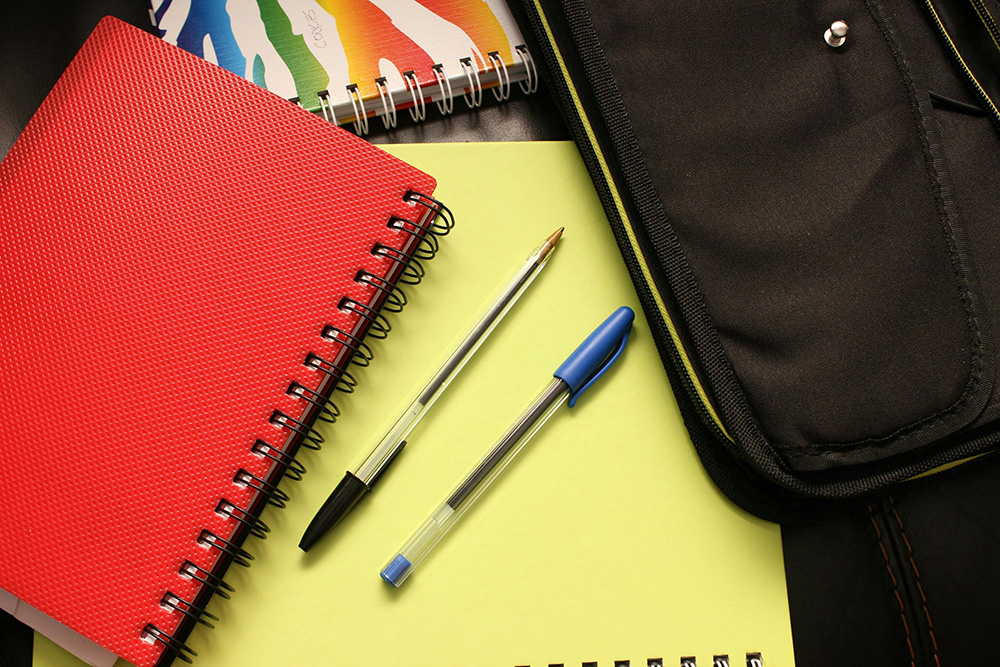 A close-up image of school supplies, including a red spiral notebook, a black-ink pen, a blue-ink pen, a yellow notebook, a multi-colored notebook, and a black pencil case—all items you might find on comprehensive supply lists—arranged on a dark surface.