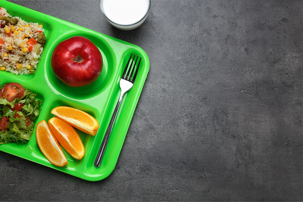 A bright green cafeteria tray holds a serving of rice with vegetables, a shiny red apple, orange slices, a fork, and a side salad with tomato pieces. A half-full glass of milk sits on the gray table next to the tray—a perfect menu for lunch.