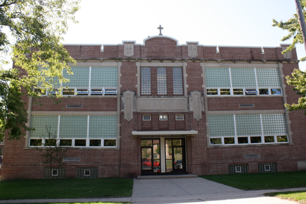 A two-story brick building with large glass block windows, an entrance in the center, and a cross at the top middle of the structure, suggesting it may be a religious or educational institution adhering to specific policies. The building is surrounded by trees and has a paved walkway leading to the entrance.