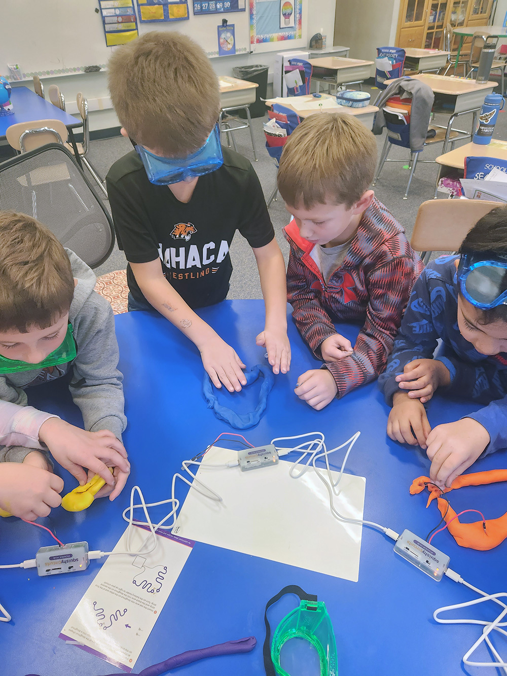 Four children are gathered around a blue table engaged in a science activity as part of an after school program. Two children are wearing safety goggles, one is touching blue clay while the others observe. Wires and small electronic devices are scattered on the table.