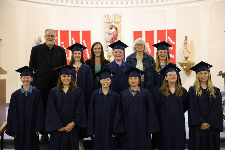 A group of 6th grade graduates in navy caps and gowns stands in two rows. Three adults, including a man in clerical attire and two women, stand behind them. The backdrop is St. Mary’s church with red and white decorations and a religious statue, marking the Class of 2024's special day.