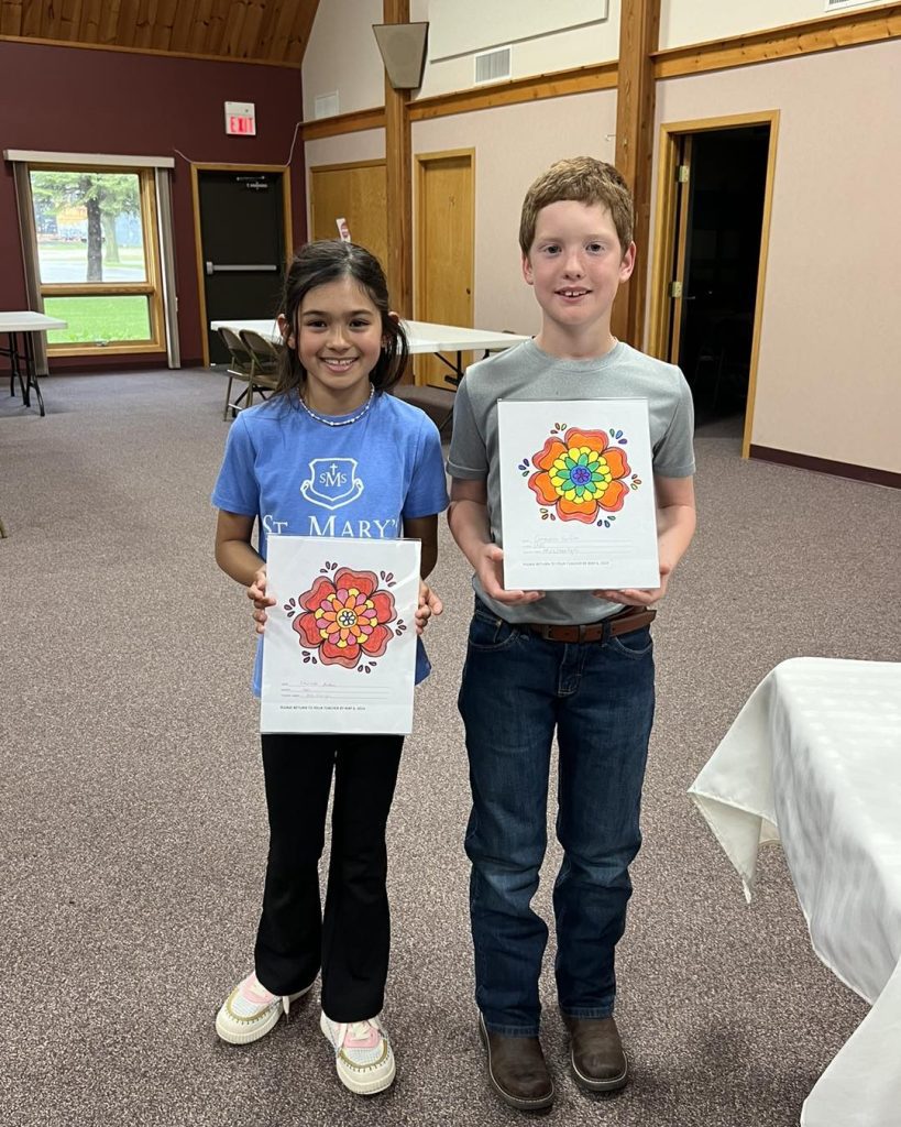 Two children stand indoors holding up their artwork. The girl on the left has long dark hair and wears a blue shirt and black pants, while the boy on the right has short red hair and wears a grey shirt and jeans. Both smile proudly as winners of the Sons of Norway coloring contest, displaying their brightly colored flower drawings.