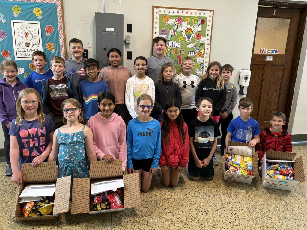 A group photo features 19 smiling school children standing in a hallway with colorful decorations on the walls. Three of the children are holding boxes filled with snacks for care packages in a donation project, as suggested by a nearby display board supporting military families.