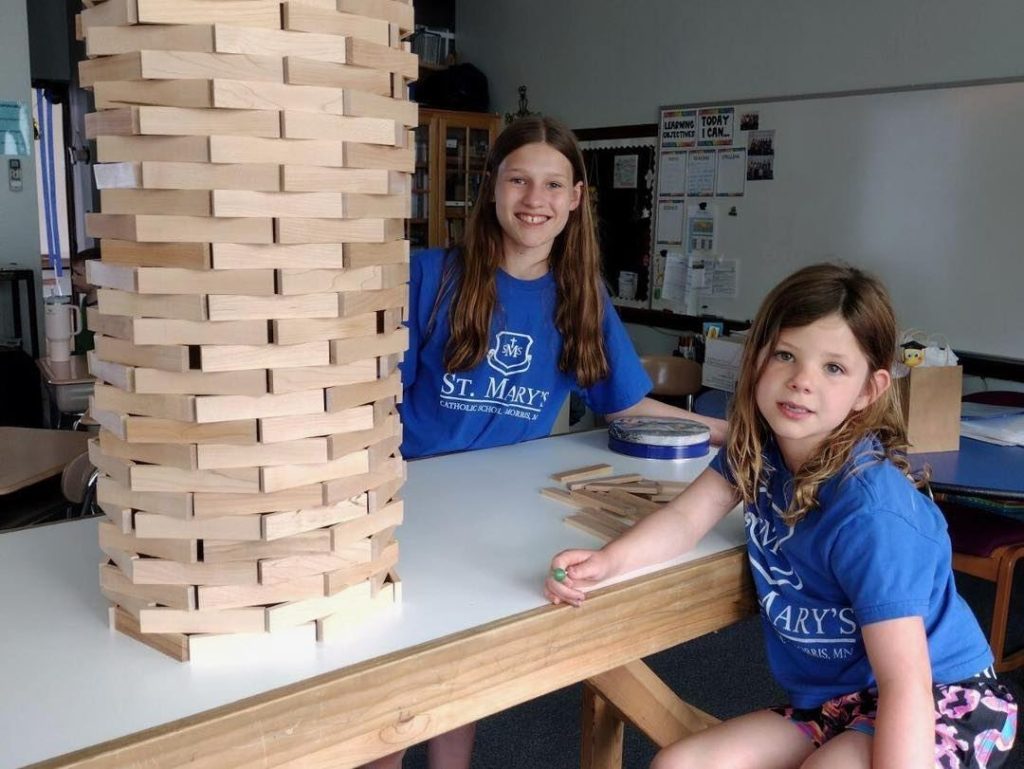 Two children, both wearing blue St. Mary's shirts, are standing beside a tall tower made of wooden blocks. The older child is smiling, and the younger child is looking towards the camera. The classroom, part of the C-STEM lending library at CSOE at the University of St. Thomas, buzzes with learning activities in the background.