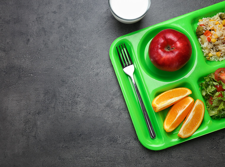 A green cafeteria tray in a Catholic school holds a fork, a red apple, orange slices, a portion of rice with corn and vegetables, and a salad with lettuce and tomatoes on a dark surface. A glass of milk is positioned near the top edge of the tray.
