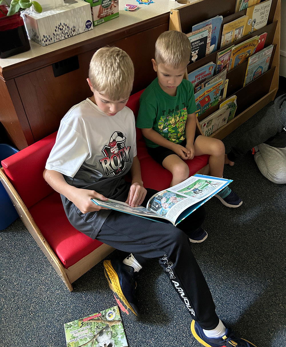 Two boys sit on a red couch at the After School Program, looking at a large picture book together. The older boy, wearing a white and black shirt and black pants, holds the book, while the younger boy in a green shirt and beige shorts attentively watches. Bookshelves are seen in the background.