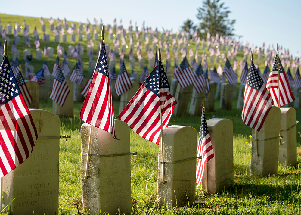 Rows of grave markers in a cemetery are adorned with American flags. The scene extends into the distance, with many flags fluttering in the breeze on a sunny day. The grass is green and well-kept, and some trees are visible in the background.