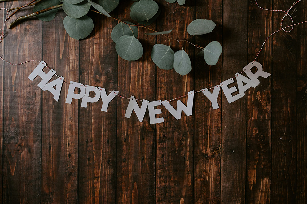 A "Happy New Year" banner is hung against a dark wooden background adorned with eucalyptus branches. The banner features white letters, creating a festive and inviting atmosphere.