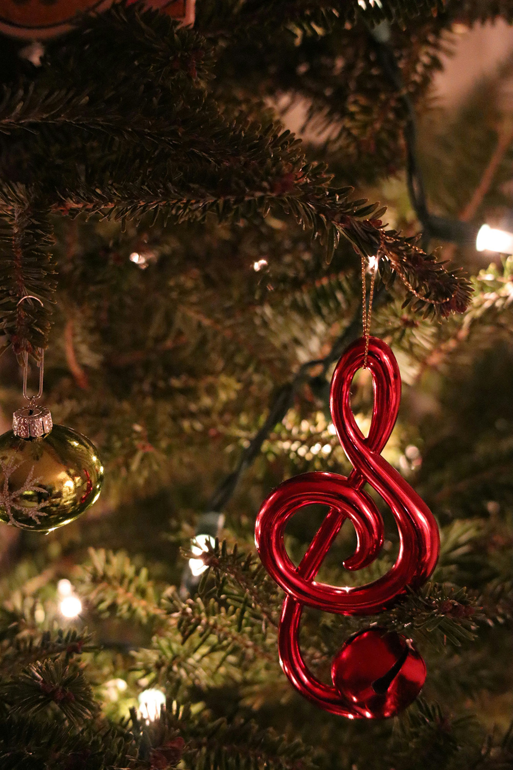 Close-up of a Christmas tree decorated with twinkling lights, a shiny green bauble, and a red treble clef ornament. The background is filled with pine needles and the warm glow of the festive lights.