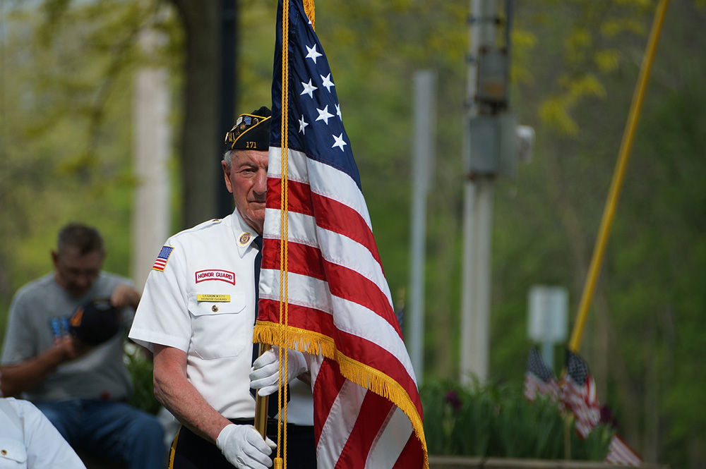 An elderly man in a uniform holds an American flag during an outdoor event. He wears a cap and white shirt with patches. Another man, blurred in the background, sits while looking down. Vegetation and additional small flags are visible in the background.