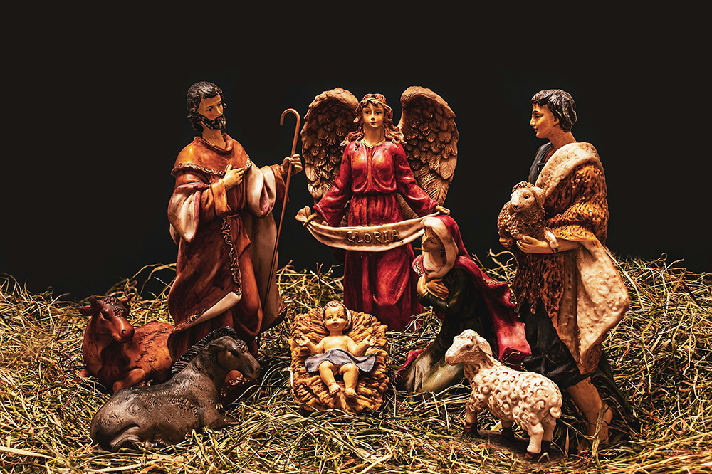 A nativity scene with figurines depicting Mary, Joseph, baby Jesus in a manger, an angel, and shepherds. Surrounding them are a cow, a donkey, and two sheep. The figures are set on a bed of straw against a dark background.