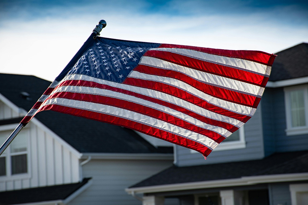 An American flag waves in the wind against a blue sky. The background features suburban houses with white and gray siding, and the rooflines and windows of the homes are visible.