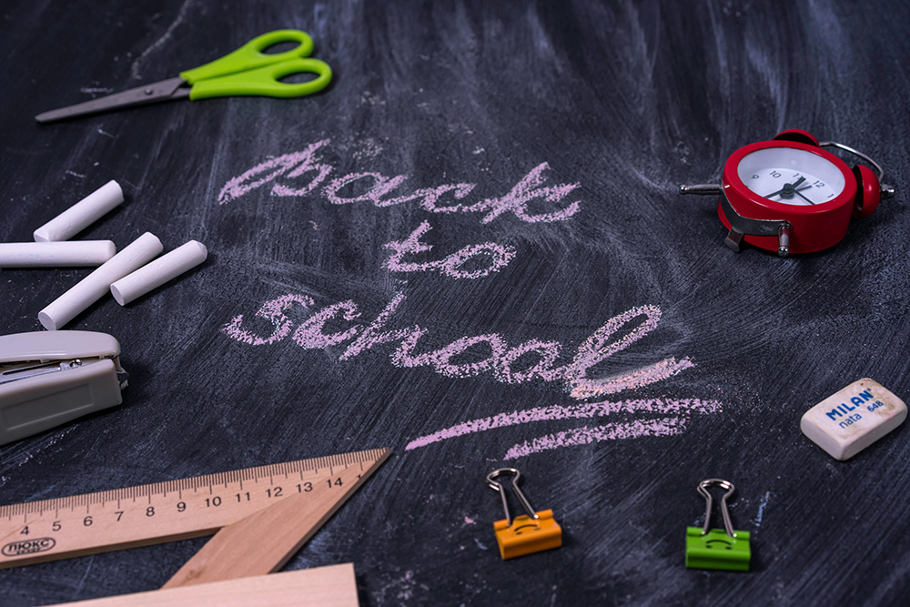 A chalkboard with "Back to School" written in pink chalk surrounded by school supplies, including green scissors, white chalk pieces, a red alarm clock, an eraser, a wooden ruler, a stapler, and colorful binder clips.