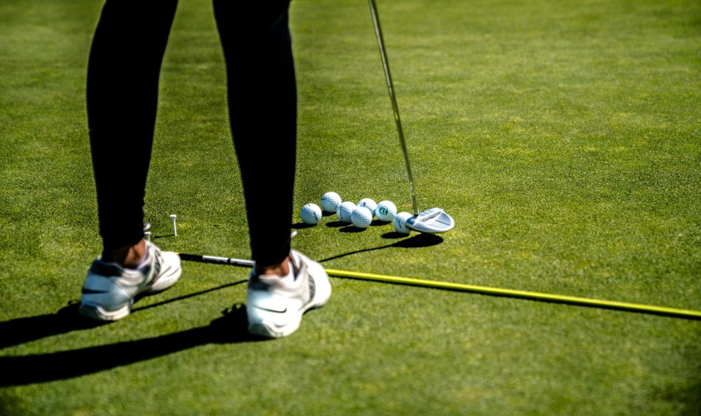 A person in white sneakers and dark pants stands on a golf course, preparing to putt a golf ball during the 4th Annual St. Mary’s Golf Tournament. Several golf balls and a golf club are scattered on the green grass, while a yellow alignment rod lies parallel to the person's feet.