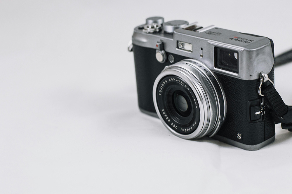 A close-up of a vintage-style camera with a sleek silver and black design. The camera has a large lens, various buttons, and dials on its top and side, and a black wrist strap attached to the right side. The background is plain white.