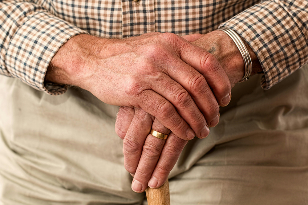 Close-up of an elderly person's hands resting on a wooden cane. The person is wearing a plaid shirt, beige pants, a gold ring on one finger, and a silver bracelet on the wrist, highlighting a sense of age and wisdom.