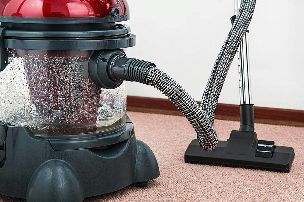 A vacuum cleaner with a transparent canister filled with dirt and dust is shown on a pink carpet. The vacuum has a red top and a black base with wheels. It is connected to a metallic hose with a handle and an attached floor nozzle, ready for some summer cleaning.