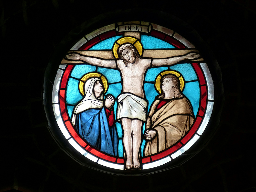 A circular stained glass window depicts Jesus on the cross with a halo, flanked by two figures, one in blue and the other in brown, both with halos, against a background of red, blue, and gold hues. The letters "INRI" are inscribed above Jesus's head.