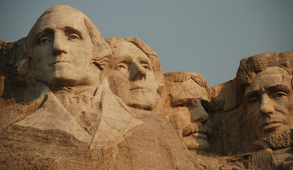 Mount Rushmore National Memorial featuring the carved faces of four U.S. Presidents: George Washington, Thomas Jefferson, Theodore Roosevelt, and Abraham Lincoln, set against a clear sky. The detailed stone sculptures are embedded in a granite cliff.