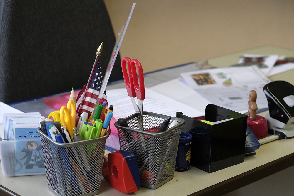 A cluttered desk with an array of office supplies, including pens, highlighters, scissors, tape, a hole punch, and a small American flag. Documents and papers are spread out on the desk in the background, along with some colorful office organizers.