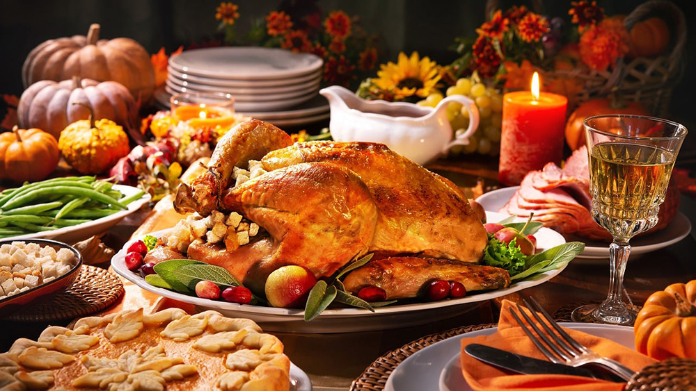 A festive Thanksgiving dinner table is set with a roasted turkey as the centerpiece, surrounded by side dishes including green beans, sliced ham, and a pie with leaf-shaped crust. There are candles, pumpkins, a glass of white wine, and a gravy boat also on the table.